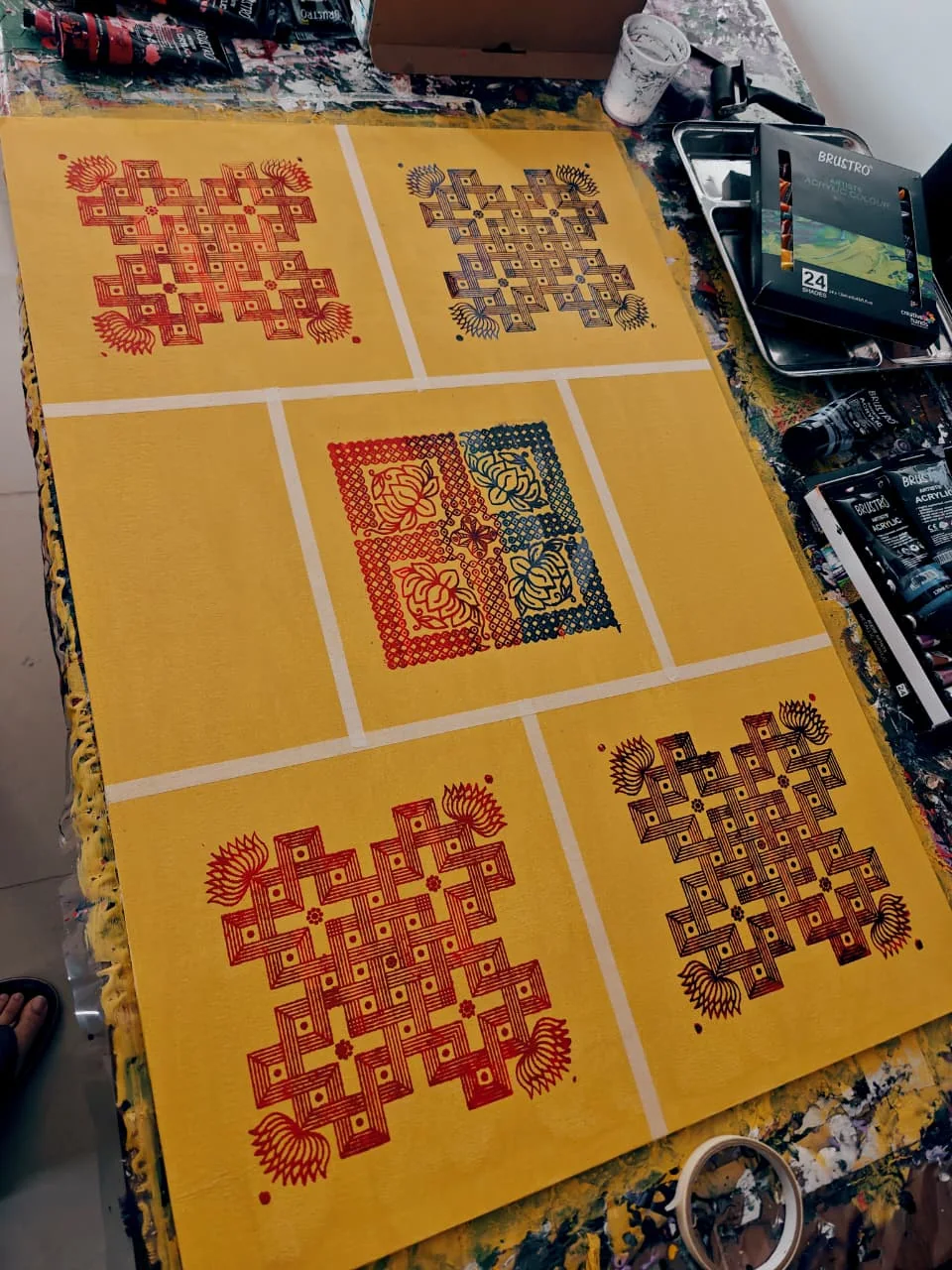 A large saffron-yellow panel photographed on a paint-spattered studio table, divided by white tape into five fields: in the four corners a square kolam-like knot of interlocking bands with lotus buds at the corners, printed in red, blue, deep red and red-brown; at the centre a square of four lotus blocks in red and blue with a lattice border; tubes of acrylic and a tin at the panel’s edge.
