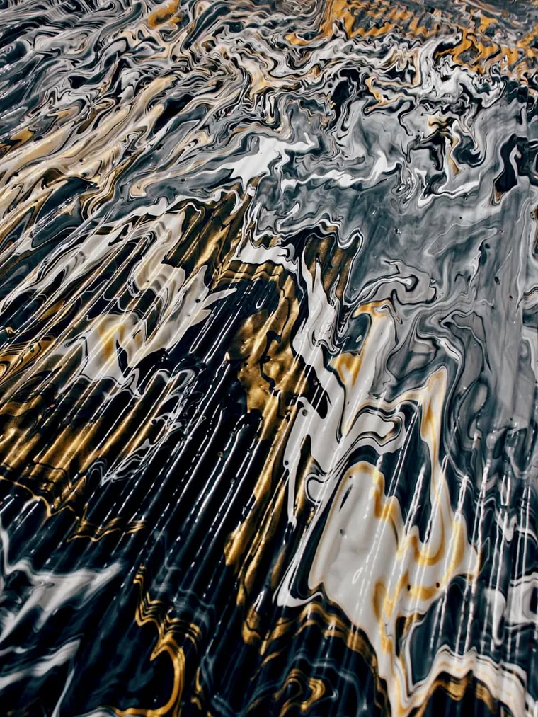 A close view of a poured painting: bands of black, grey-white and metallic gold run in long vertical drips down the surface, breaking into feathered swirls at the top where the colours meet, glossy like wet stone.