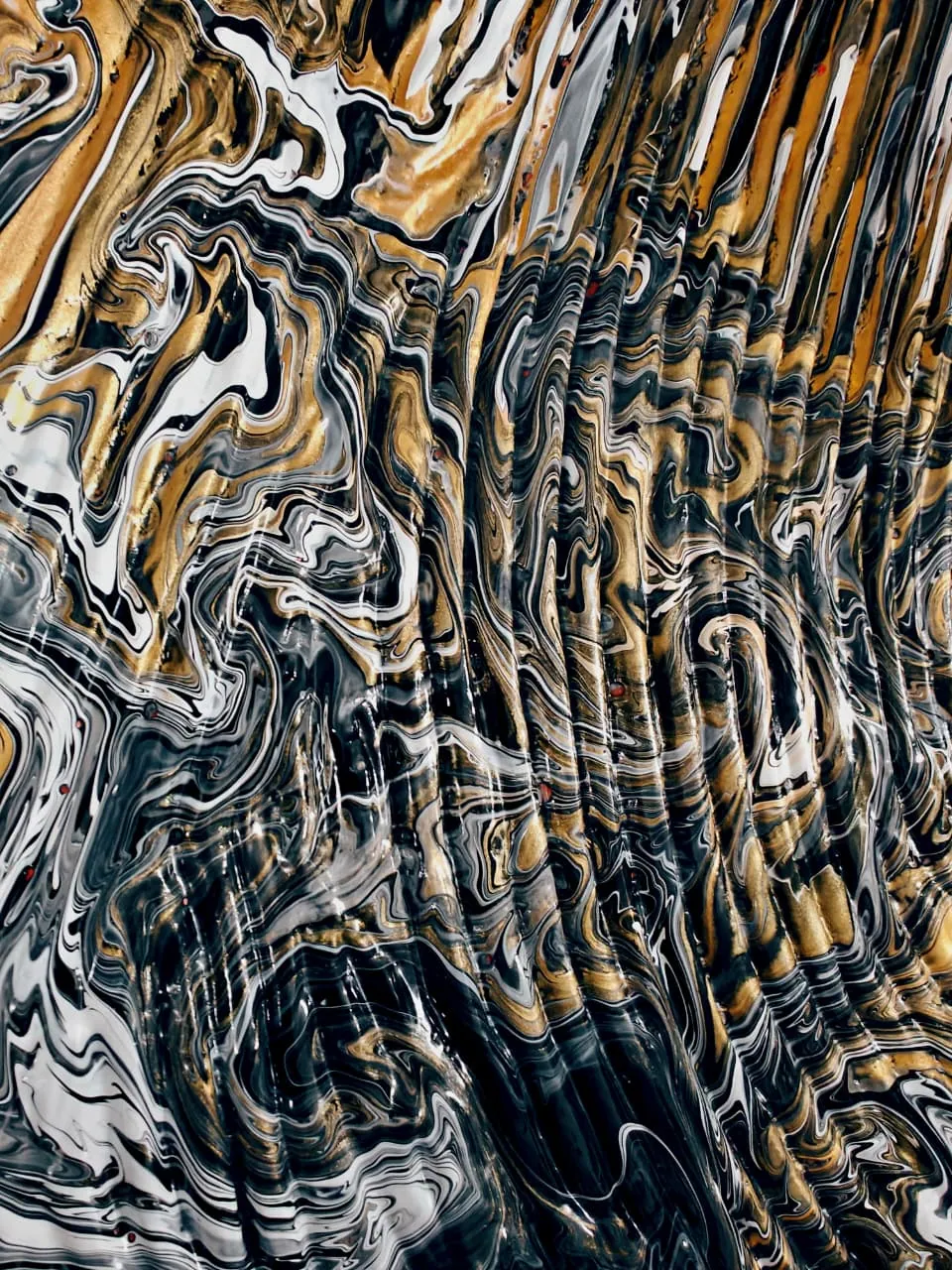 A close view of a poured painting dominated by metallic gold: broad gold bands and eddies across the upper half, folding down into black and white marbling below, the surface rippled like a seam of ore in rock.